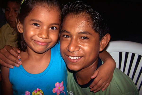 NPH Bolivia Young girl with dark shoulder length hair and an older boy with their arms around one another smiling at the camera.