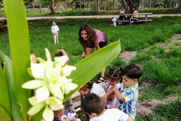 Mujer joven inclinada con las manos sobre las rodillas mirando a un grupo de niños jugando con una flor en primer plano.